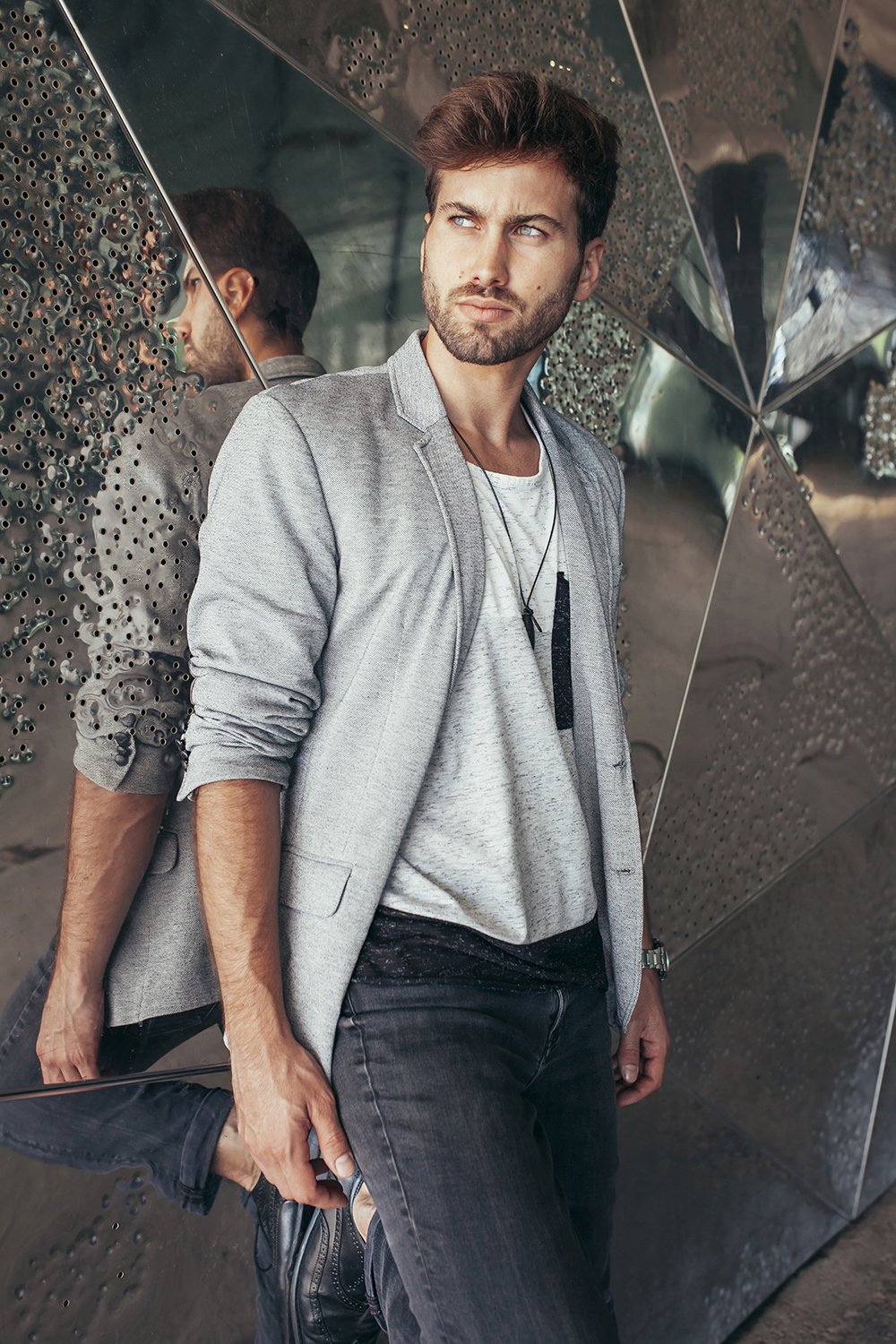Fashionable handsome man in stylish gray jacket and casual t shirt standing and looking away with reflected modern wall on background.