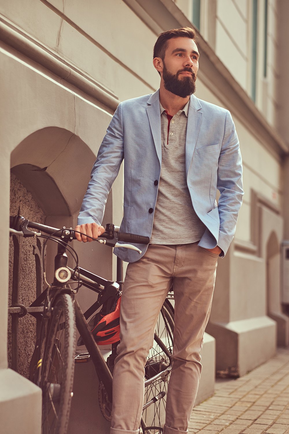 Fashionable bearded male wearing stylish clothes, stands with a bicycle on an antique street in Europe.