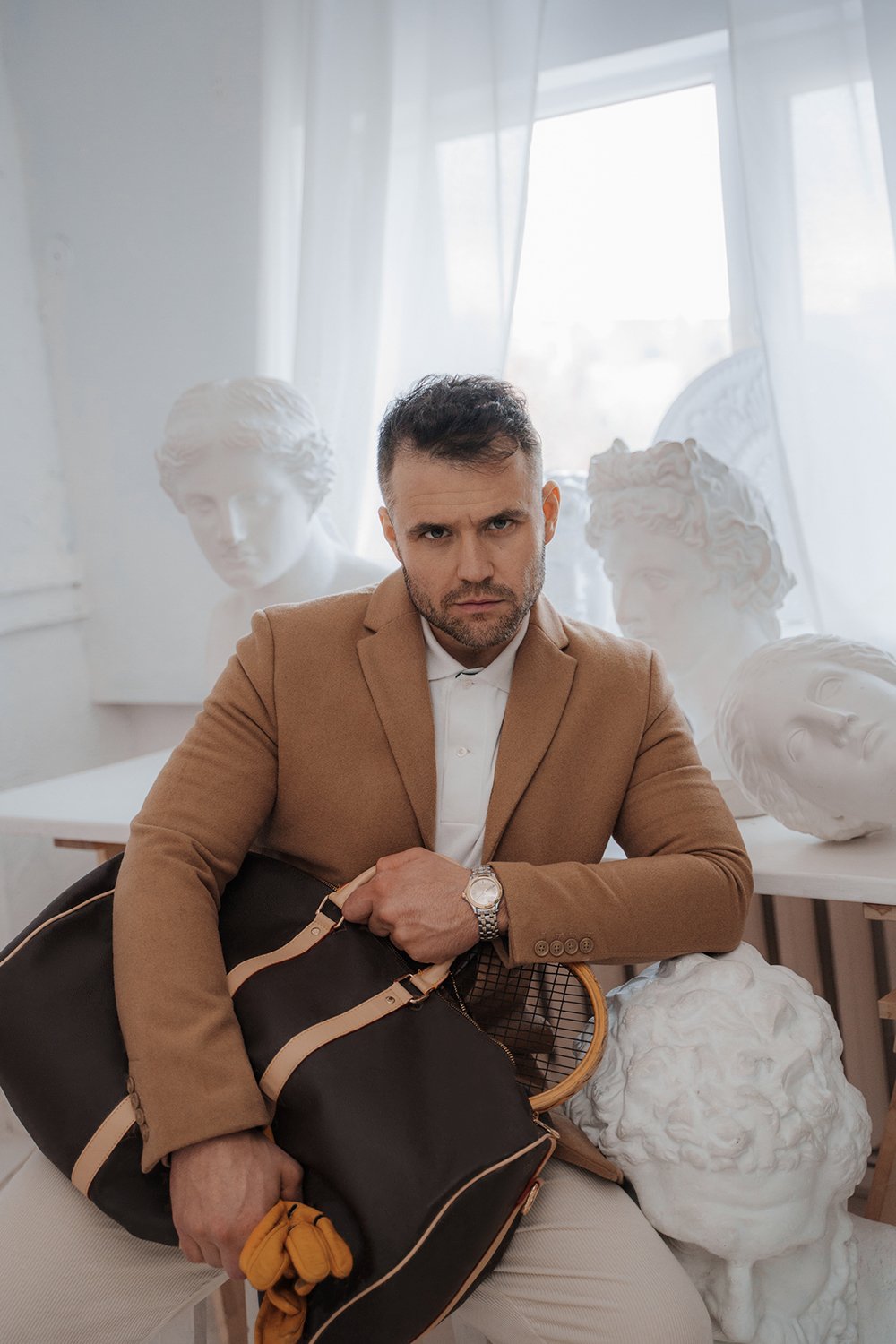 Wealthy man in old money style holding a bag and tennis racket in sculptor's studio