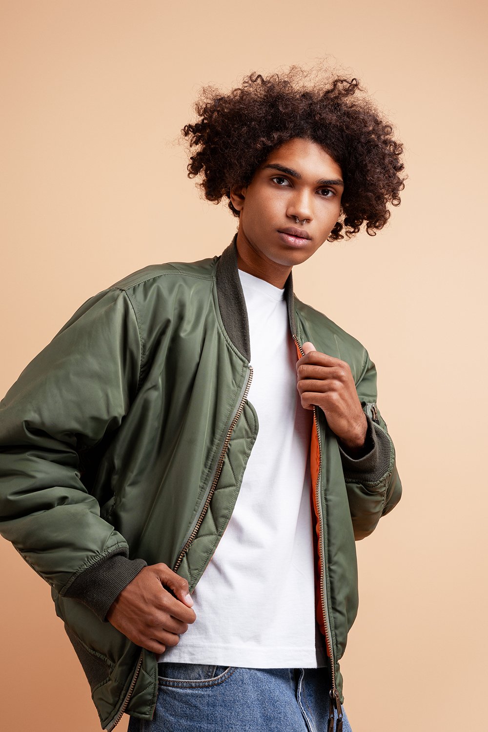 Pensive, fashion African American young man with piercing in nose wearing stylish green jacket looking at camera posing isolated on beige background. Advertisement concept